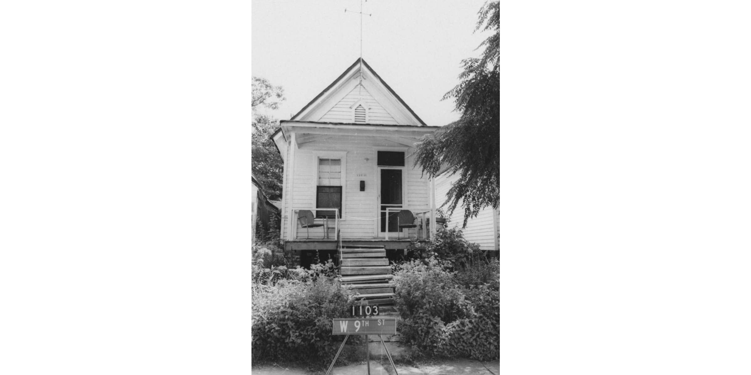 A black and white photograph of the front of a house with chairs on the porch. 