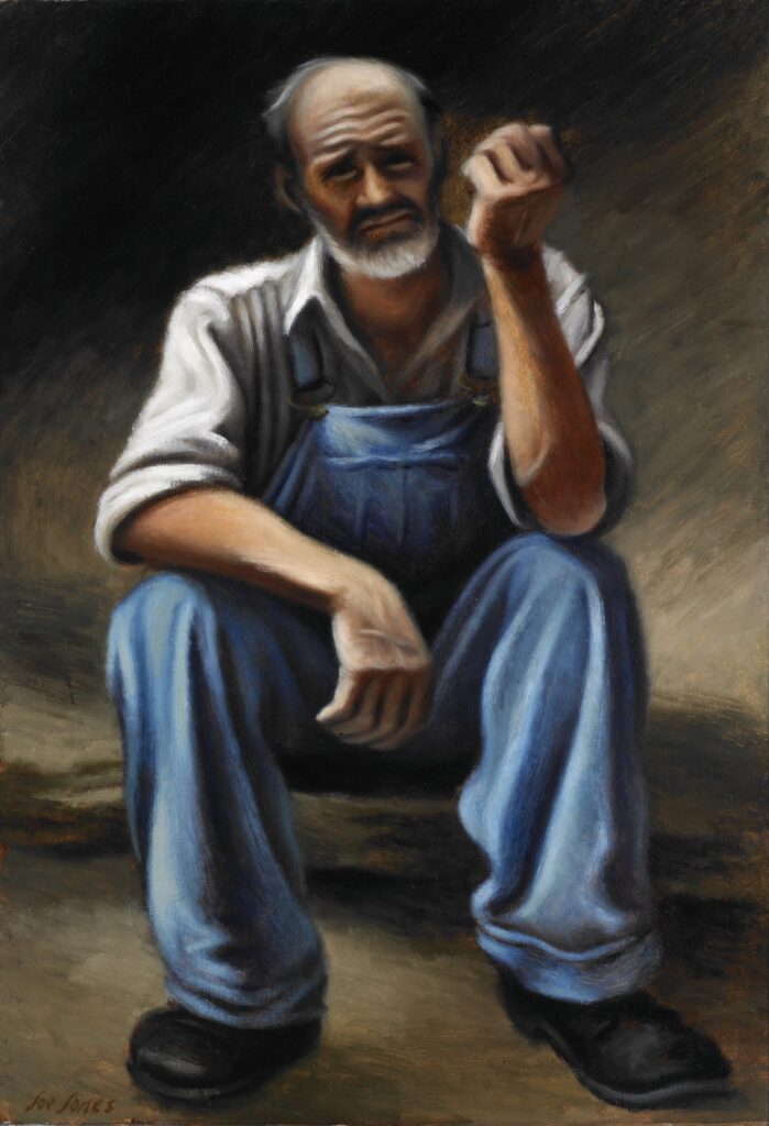 A man in overalls sits on the grond with his arms on his knees, looking straught.