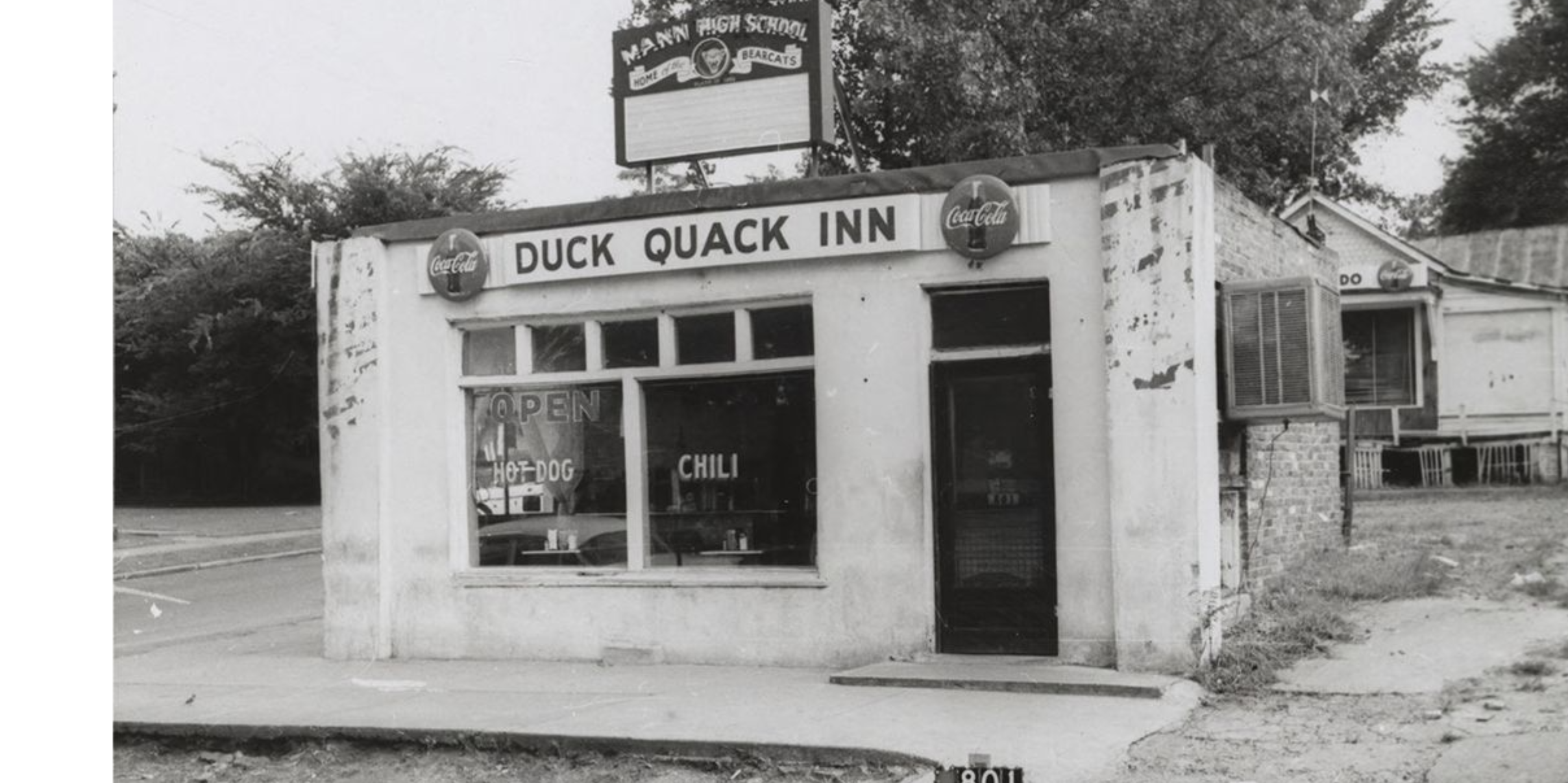 Exterior of a small building with a sign that reads "Duck Quack Inn." Signs in the front windows advertise hot dogs and chili.