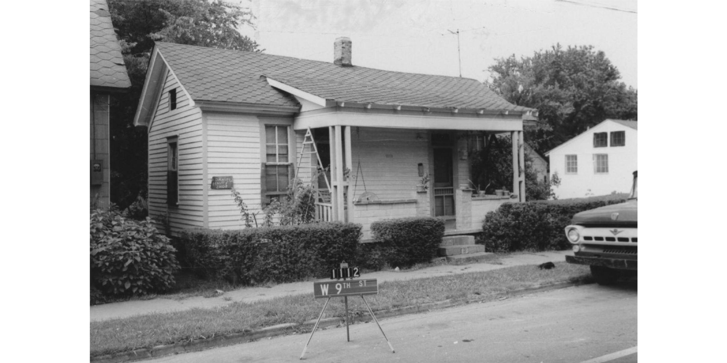 A small house surrounded by shrubs. A small, hand-written sign on the left reads "Daniels [?] Beauty Shop."