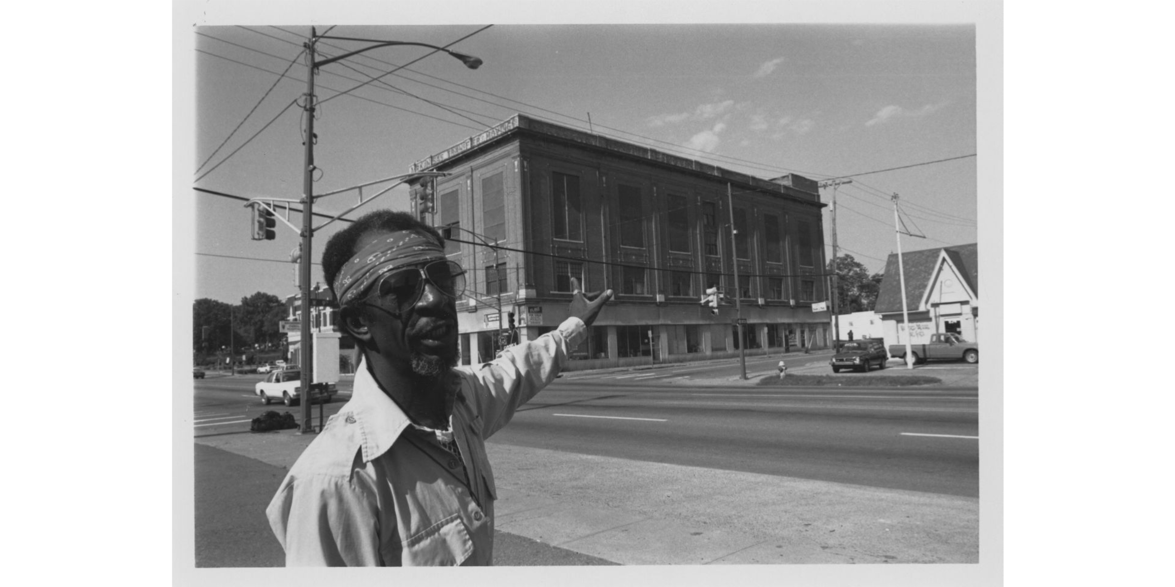 A man in a button up shirt, sunglasses, and a bandana, gestures towards a multi-story brick building.
