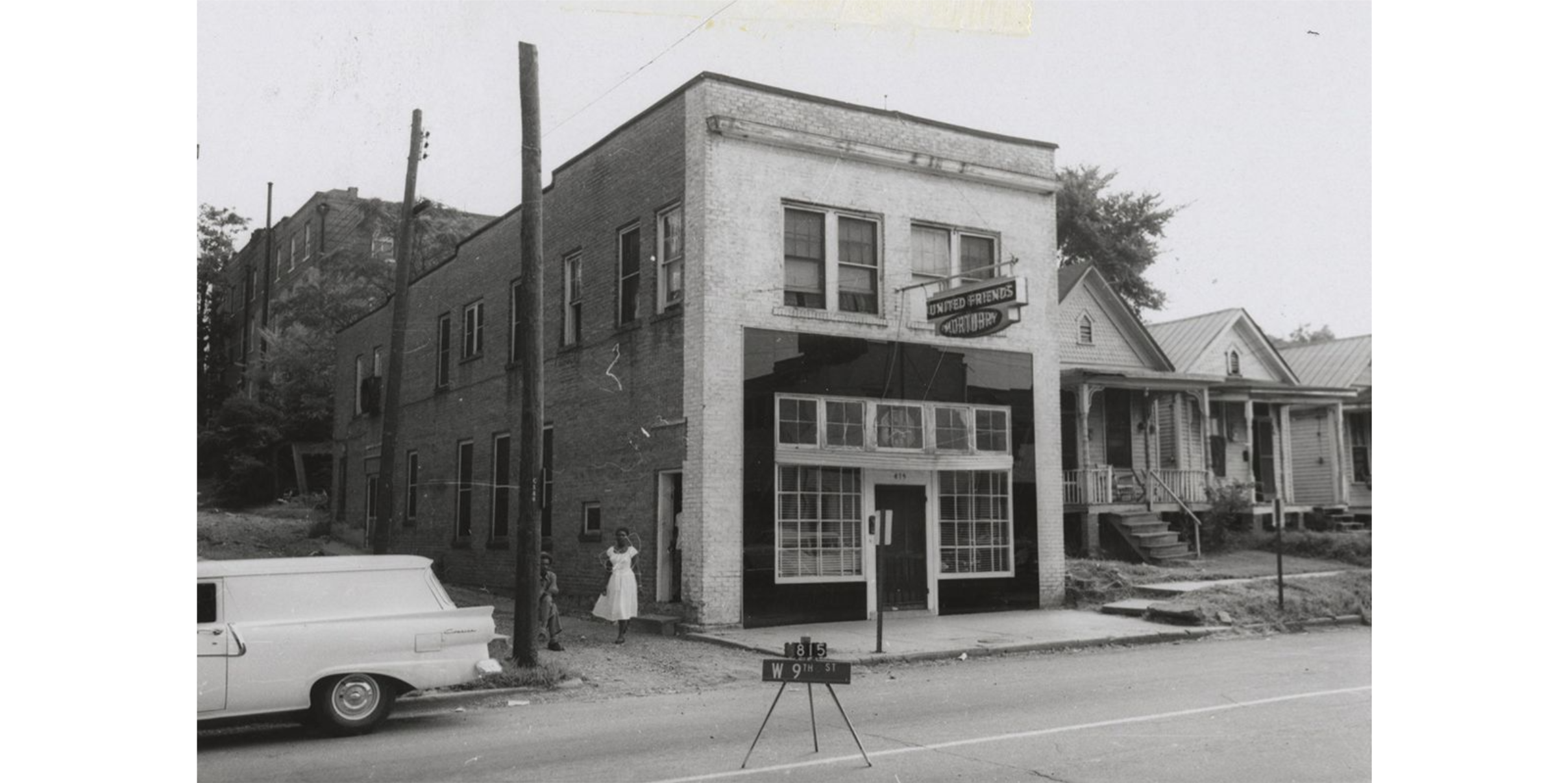 Two-story brick building with a sign which reads "United Friends Mortuary" hanging above the door. A woman stands near the side entrance on the left side of the building.