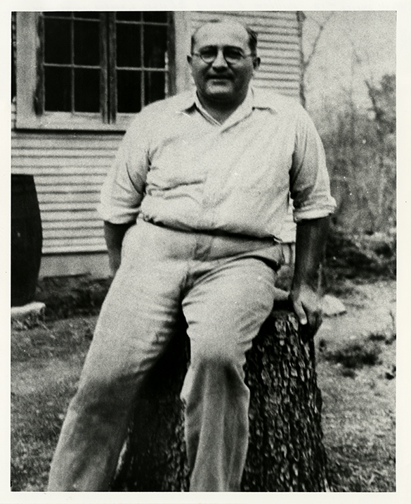 A man in glasses and a button down shirt sitting on a tree stump. 