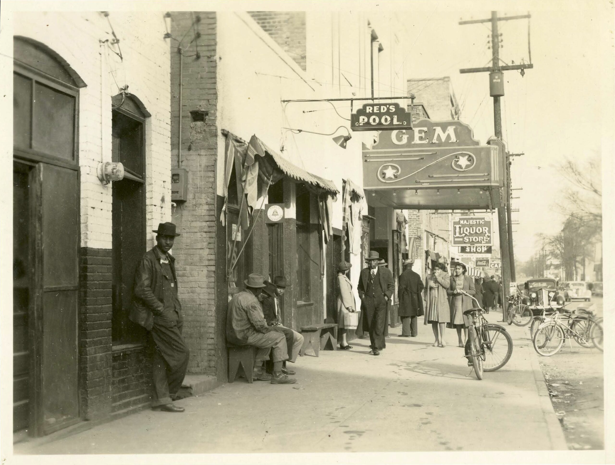 African American men and women walking along a busy sidewalk lined with businesses. The visible businesses in this photo include Red's Pool, Gem Theater, and Majestic Liquor Store.
