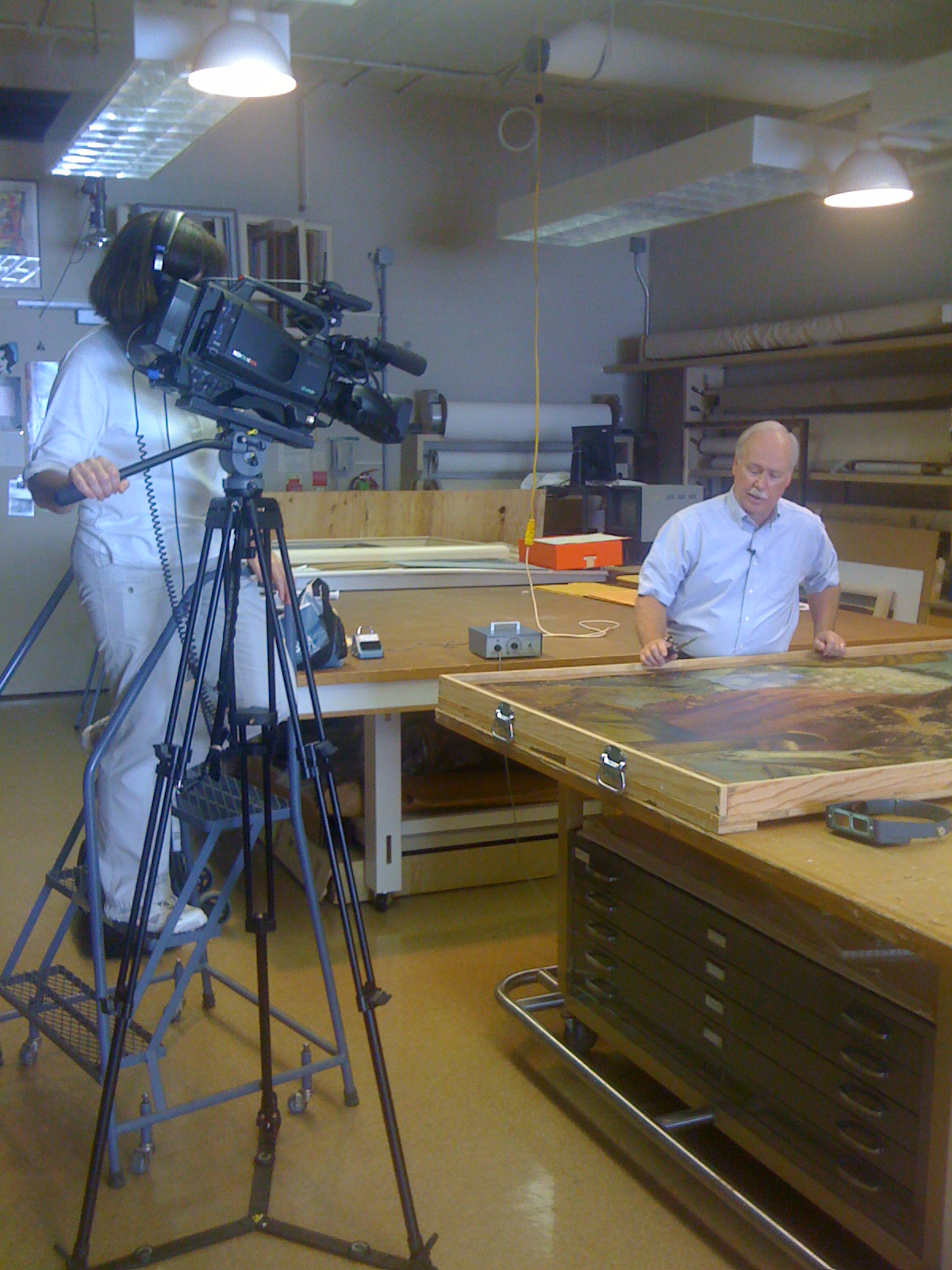 A man standing next to a large table with a large painted panel laying flat on the surface. To the left, a person with a professional camera is filming.