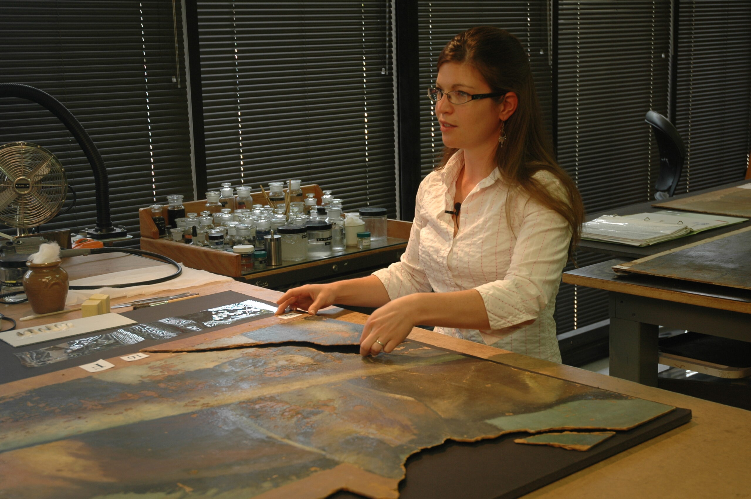 A woman standing behind a table, holding a fragmented piece of art. On the table's surface, there is a large piece of fragmented art.