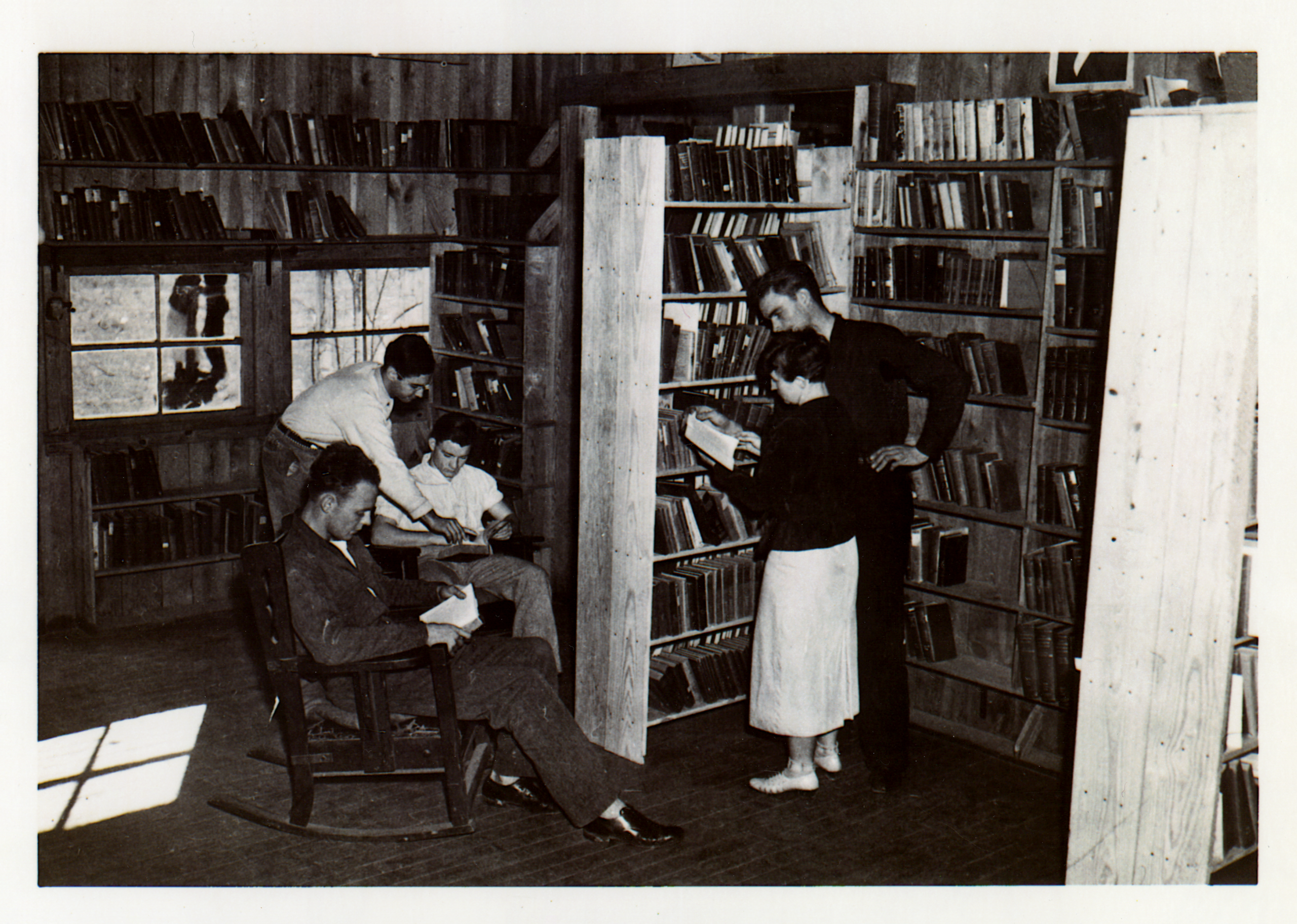 Five people in a library. Two people are sitting in rocking chairs with books in their hands.
